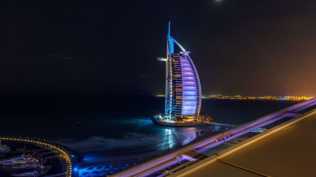 Nighttime view of the marsa al araba| Burj Al Arab hotel in Dubai, illuminated in blue and purple hues, standing on its own artificial island with the Arabian Gulf surrounding it. The distant city lights and marina with yachts add to the luxurious ambiance.