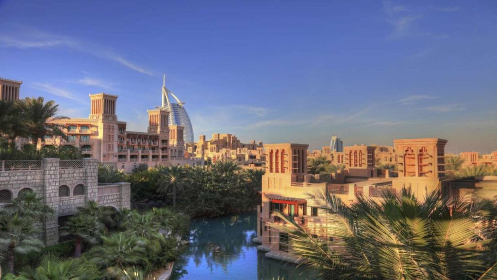 Scenic view of Madinat Jumeirah in Dubai featuring traditional Arabian-style architecture surrounded by lush greenery and waterways, with the iconic Burj Al Arab hotel prominently visible in the background under a clear blue sky.